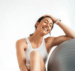 A woman in workout clothes sitting and smiling leaning into a workout ball