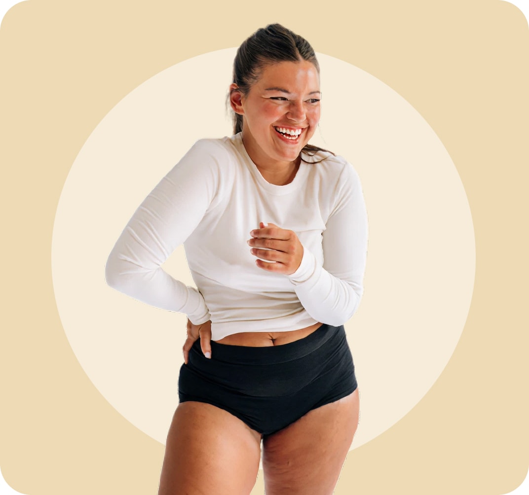 Woman in a white long-sleeve top and black high-waisted underwear, laughing and looking to her left with one hand on her waist and the other across her stomach.